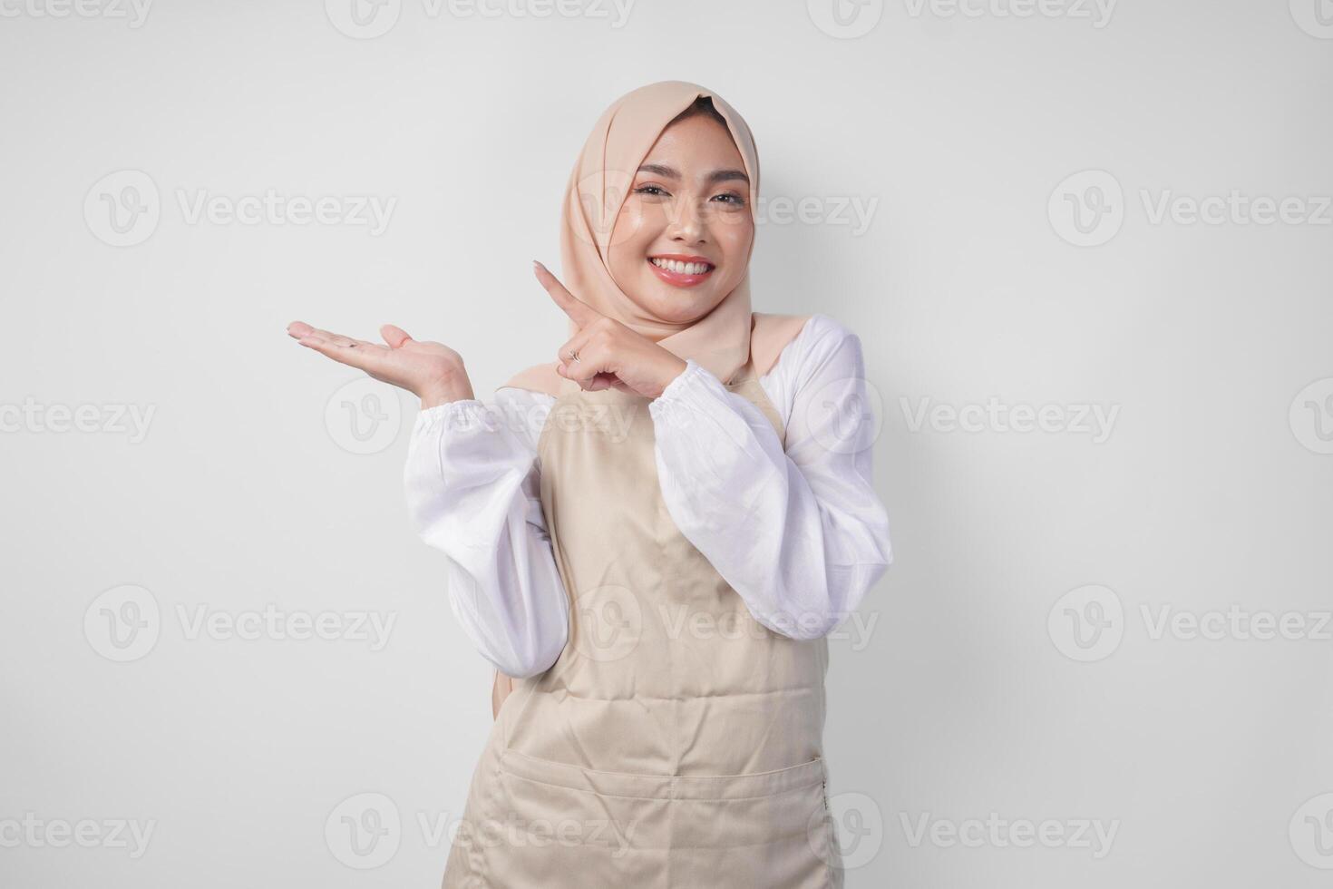 Excited young Asian Muslim woman in hijab and cream apron pointing to the empty copy space on the left and right side. Ramadan advertisement concept photo