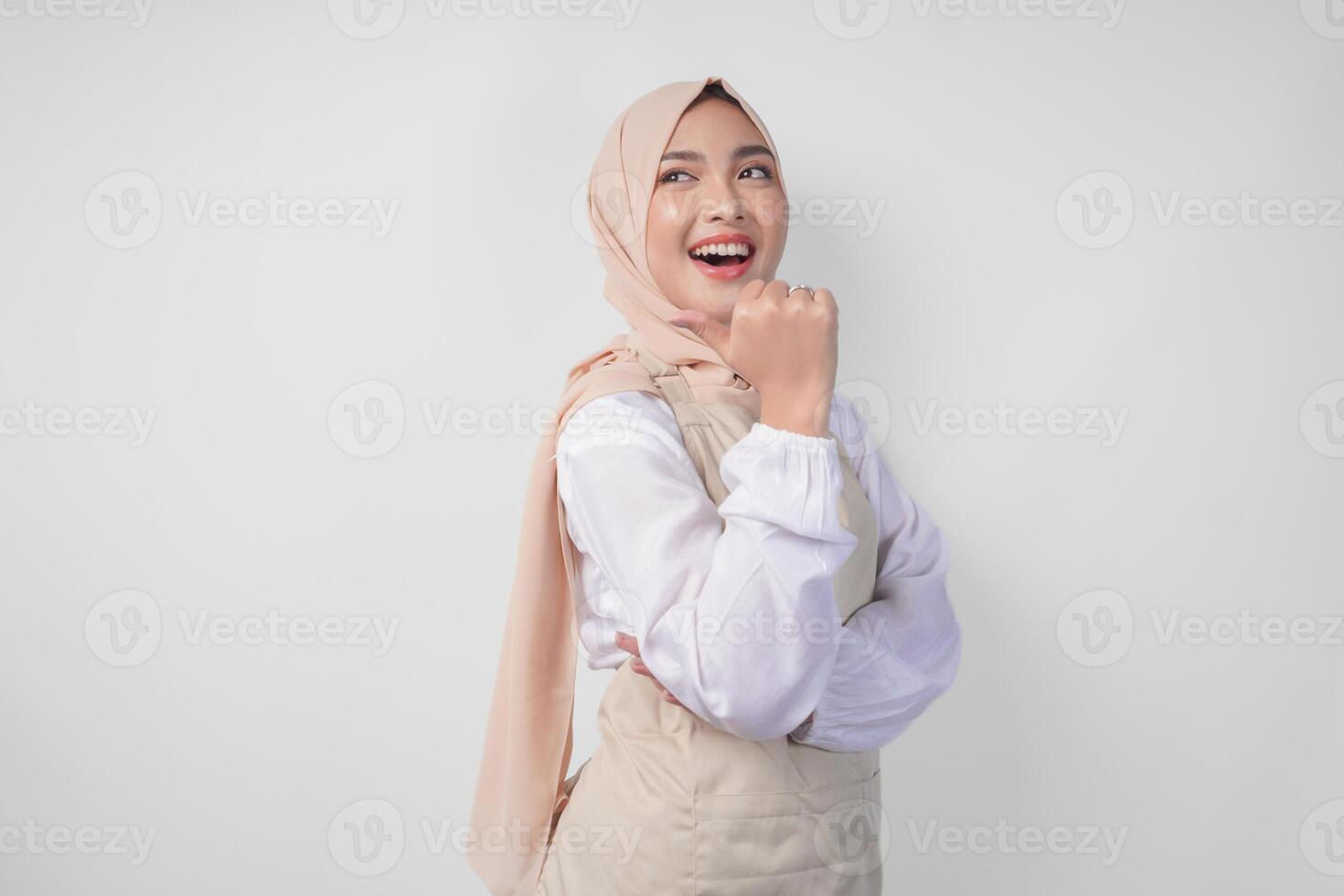 Excited young Asian Muslim woman in hijab and cream apron pointing to the empty copy space on the left and right side. Ramadan advertisement concept photo