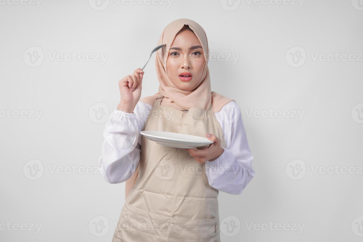 Confused young Asian Muslim woman in hijab and cream apron holding spoon and empty plate with copy space over it, thinking hard what food menu to eat for iftar. Ramadan concept photo