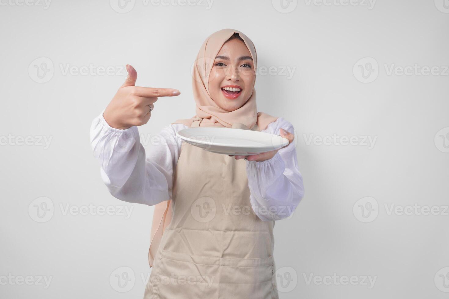Portrait of a cheerful young Asian Muslim woman in a veil hijab and cream apron pointing to an empty plate with copy space over it, presenting food menu photo