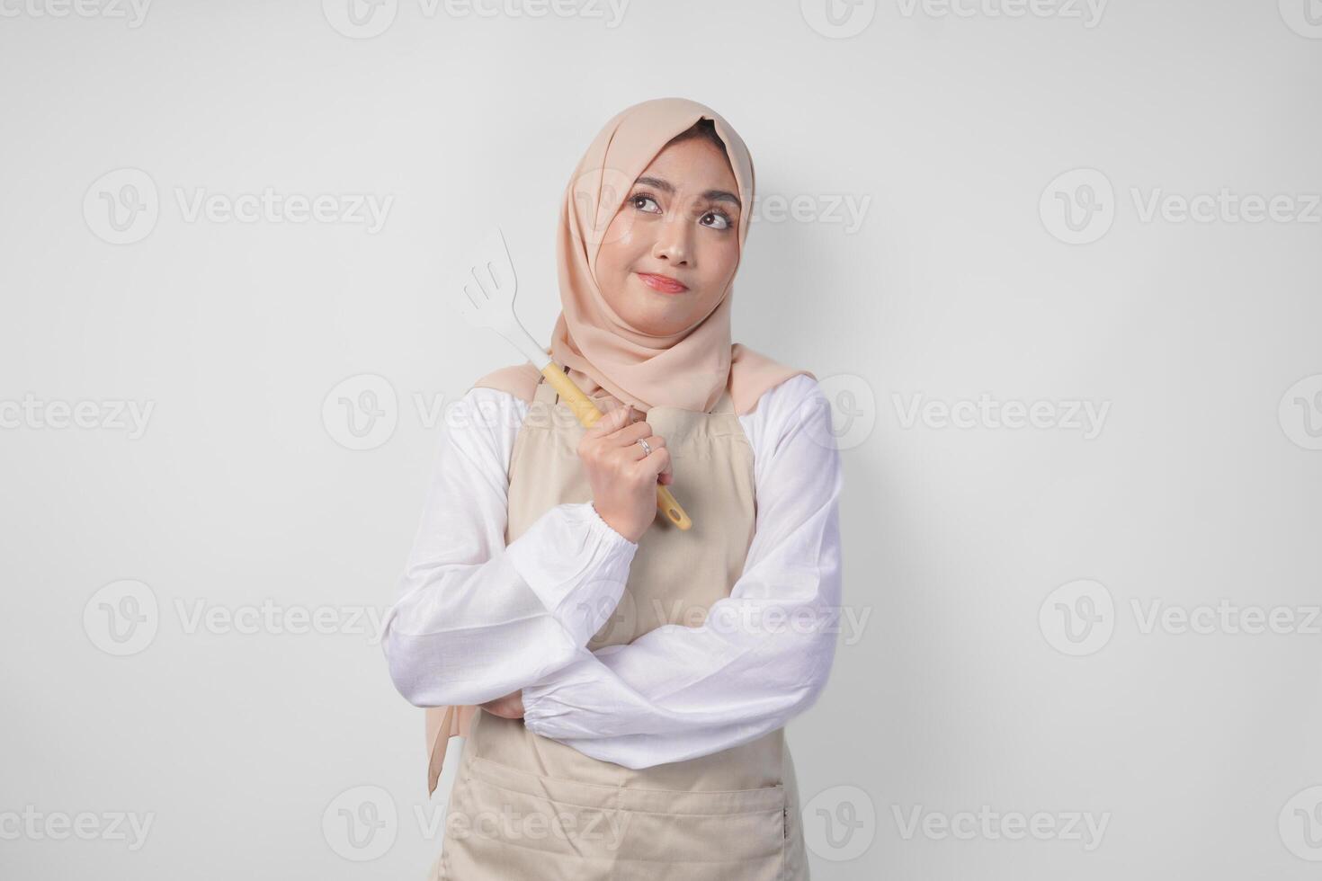 Thoughtful young Asian Muslim woman in hijab and cream apron holding spatula and kitchen cooking utensils, thinking hard what food menu to cook photo