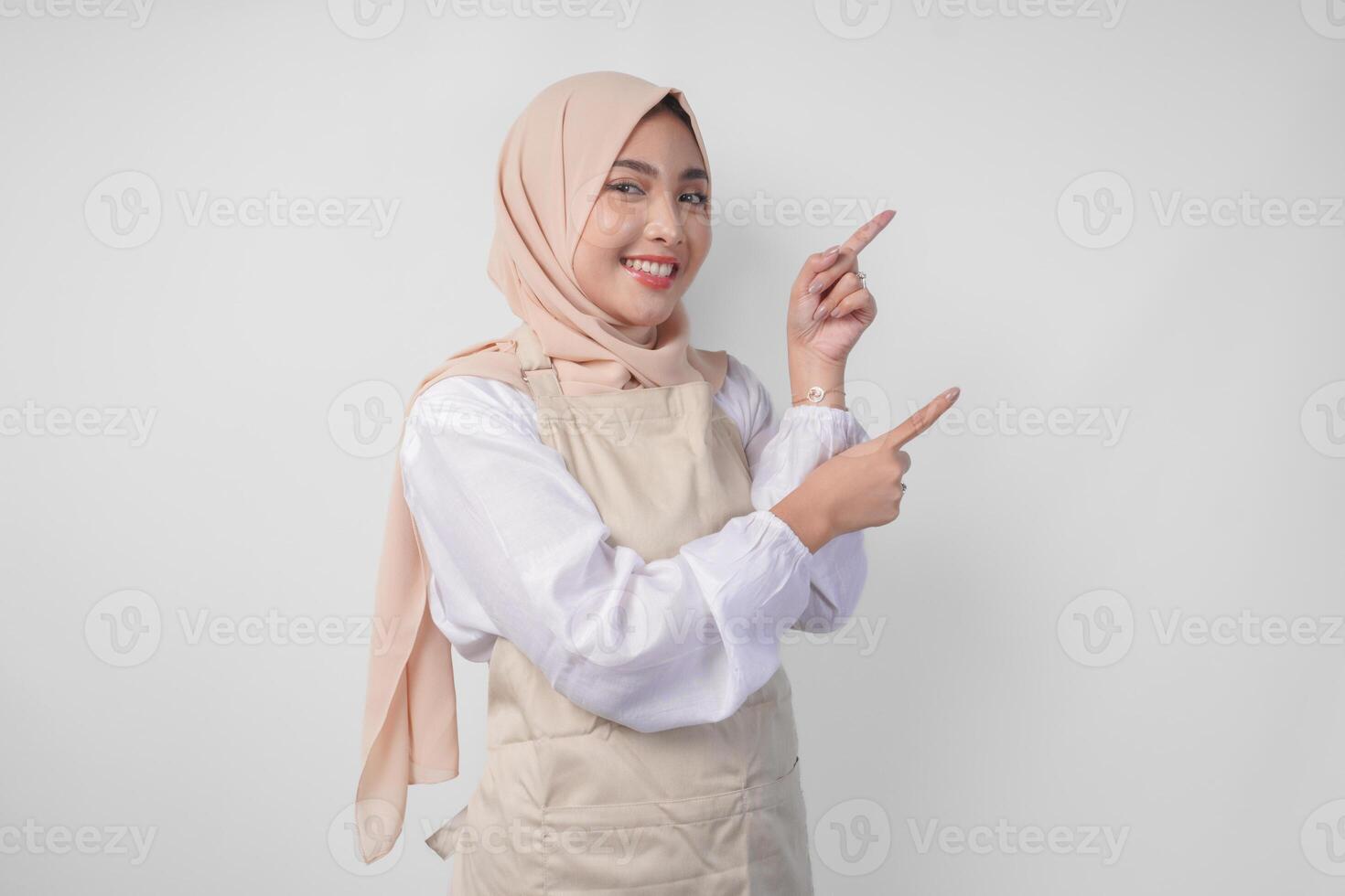 Excited young Asian Muslim woman in hijab and cream apron pointing to the empty copy space on the left and right side. Ramadan advertisement concept photo