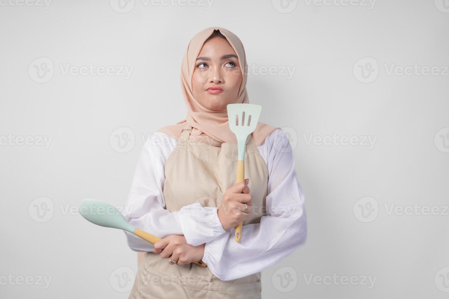 Thoughtful young Asian Muslim woman in hijab and cream apron holding spatula and kitchen cooking utensils, thinking hard what food menu to cook photo