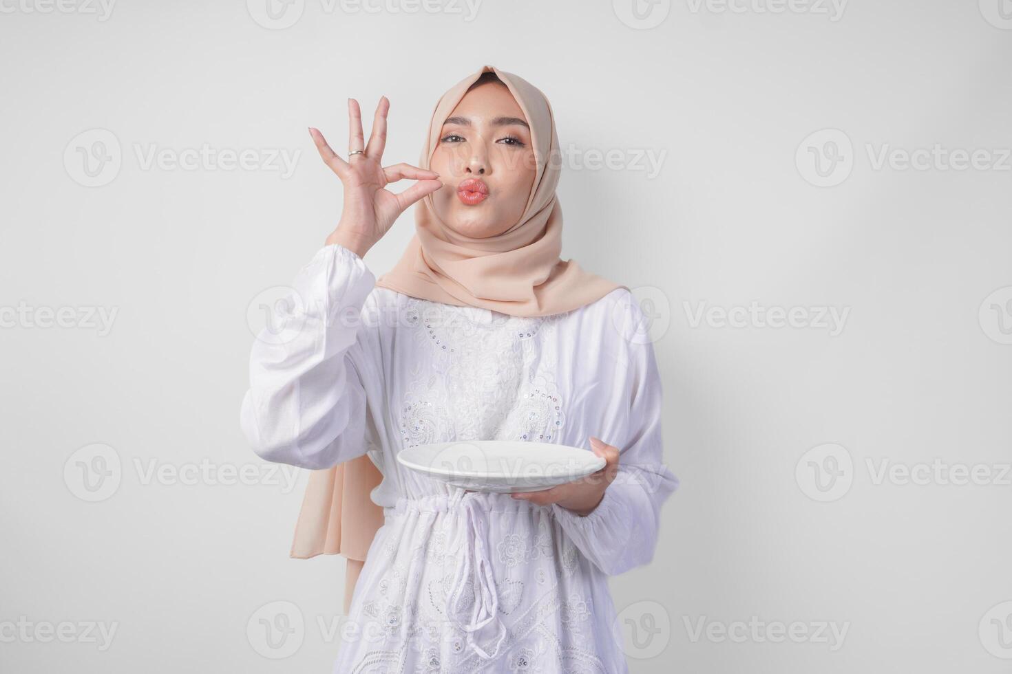 Happy young Asian Muslim woman in hijab holding an empty plate with copy space and making a delicious hand gesture to express how good the food is photo