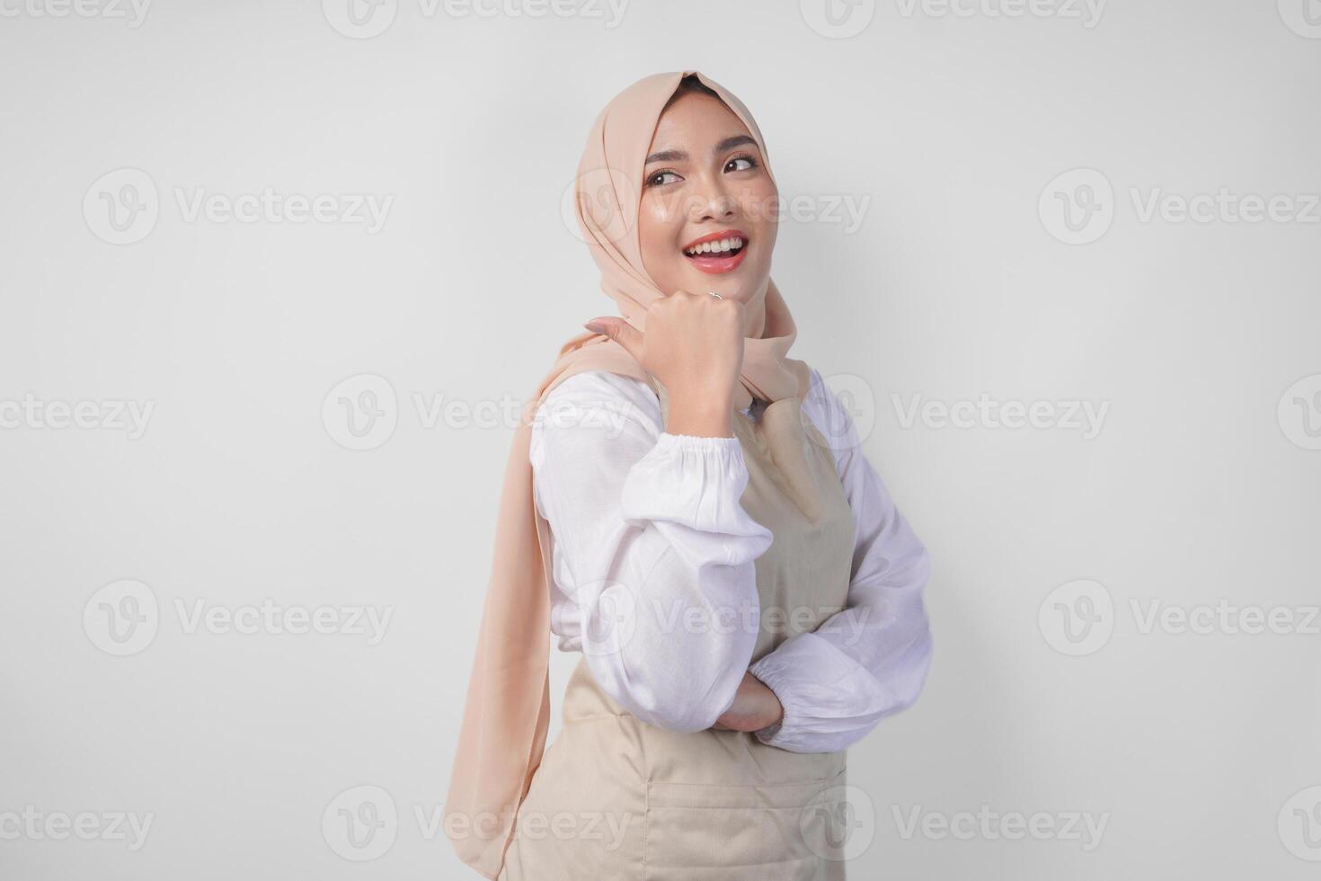 Excited young Asian Muslim woman in hijab and cream apron pointing to the empty copy space on the left and right side. Ramadan advertisement concept photo