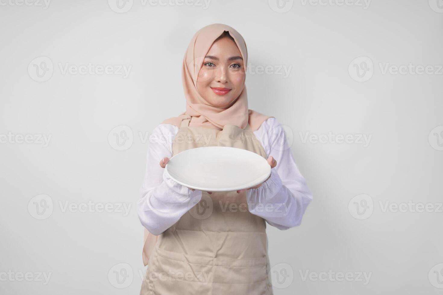 hermosa joven asiático musulmán mujer en hijab y crema delantal presentación un vacío plato con Copiar espacio terminado él, demostración el comida menú para iftar foto