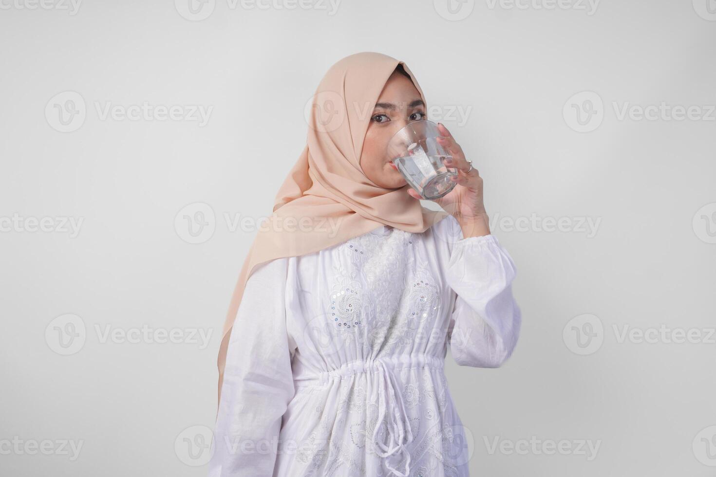 Thirsty young Asian Muslim woman in white dress and cream veil hijab drinking fresh mineral water after fasting. Ramadan concept photo