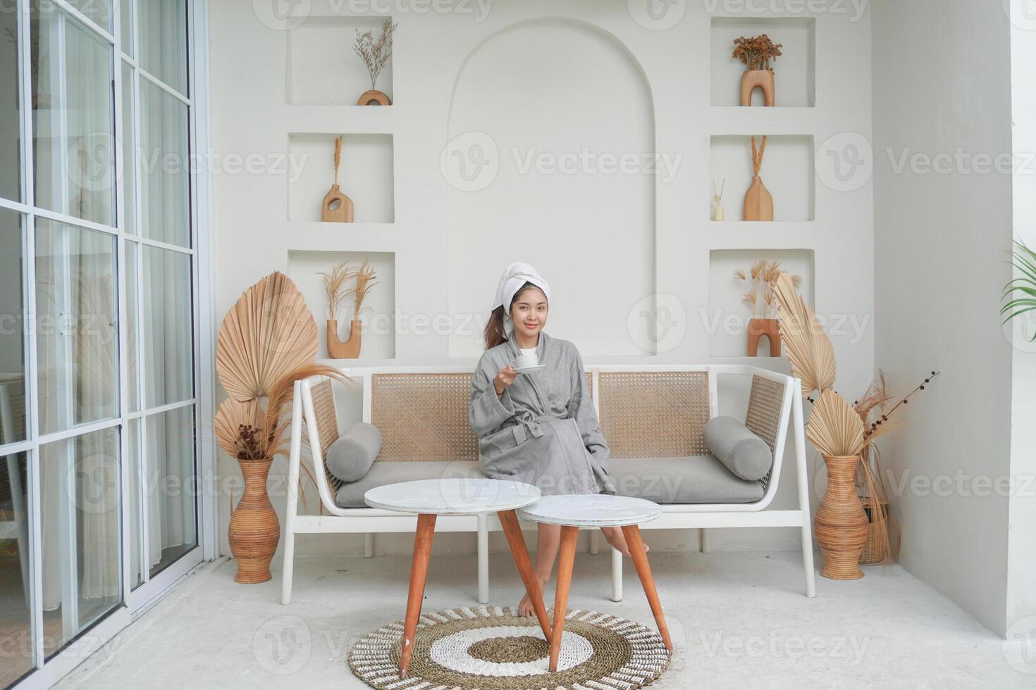 Happy attractive Asian woman in grey bathrobe posing at the terrace, smiling at camera while drinking tea. Holiday leisure concept. photo