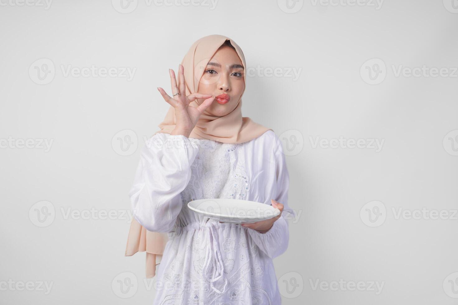 Happy young Asian Muslim woman in hijab holding an empty plate with copy space and making a delicious hand gesture to express how good the food is photo