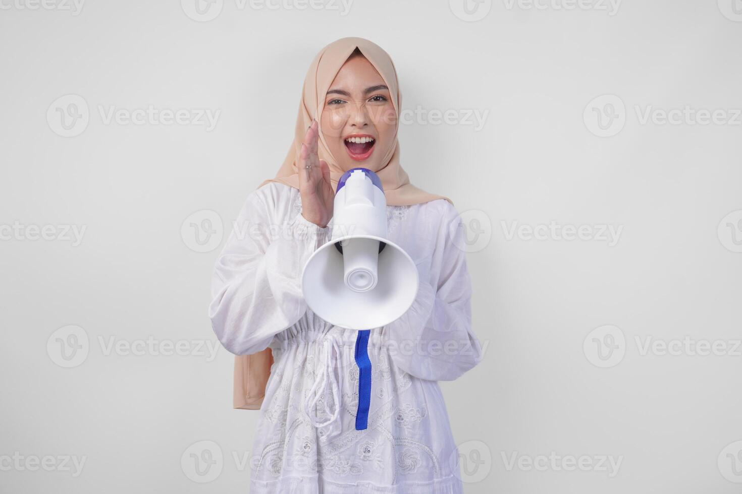 Overjoyed Asian woman wearing hijab and white dress shouting to the megaphone making announcement, standing over isolated white background photo