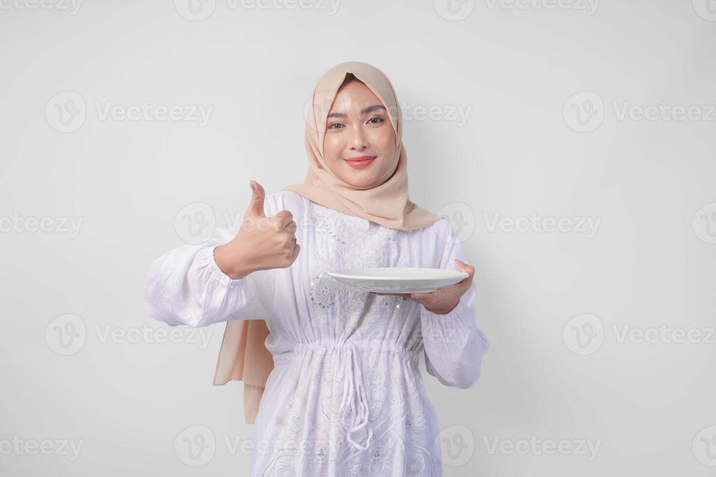 Excited young Asian Muslim woman in hijab presenting empty white plate copy space while doing thumbs up gesture photo