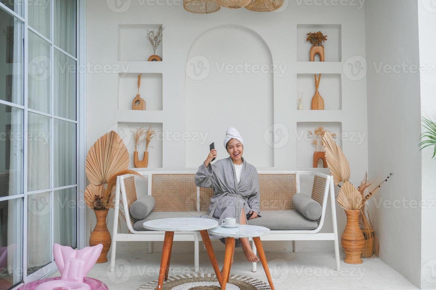 An ecstatic successful Asian woman in grey bathrobe using the smart phone sitting on the couch, lifestyle and leisure concept photo