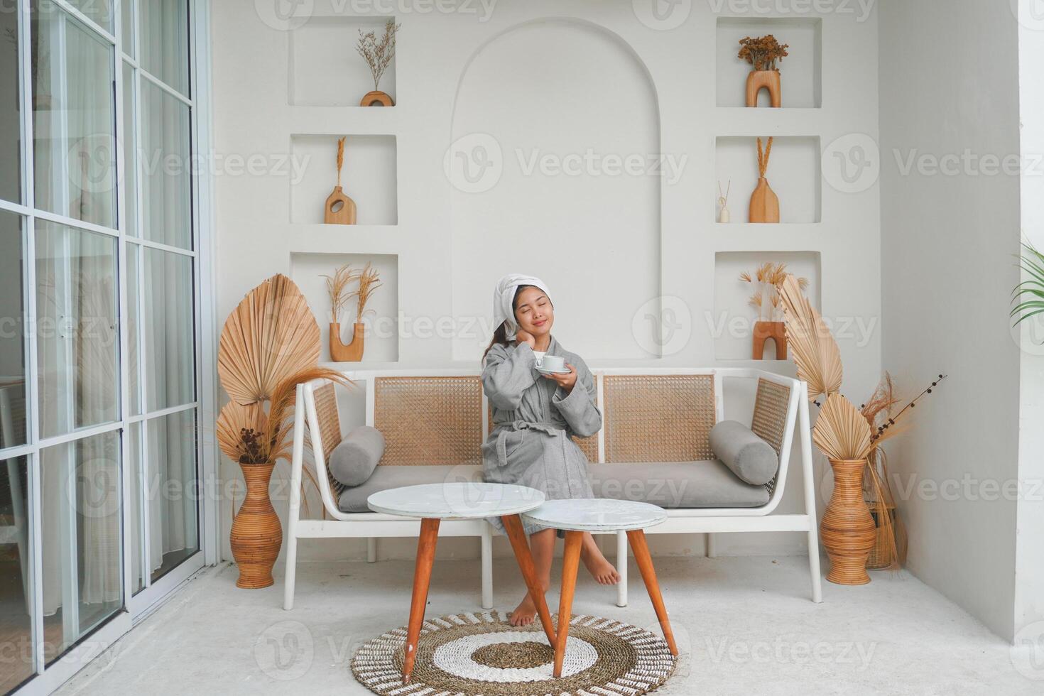 atractivo asiático mujer en gris bata de baño posando a el terraza, sonriente y gesticulando alivio o relajarse actitud mientras Bebiendo té. fiesta ocio concepto. foto