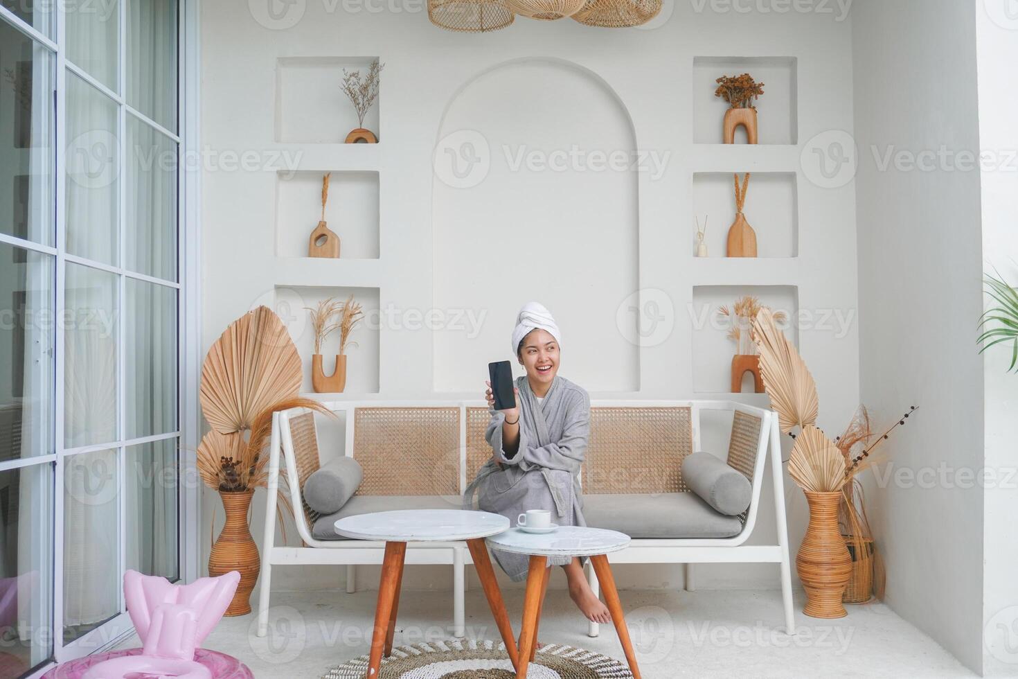 contento asiático mujer vistiendo gris bata de baño sentado en un sofá, demostración inteligente teléfono pantalla mientras sonriente alegremente fiesta ocio concepto. foto