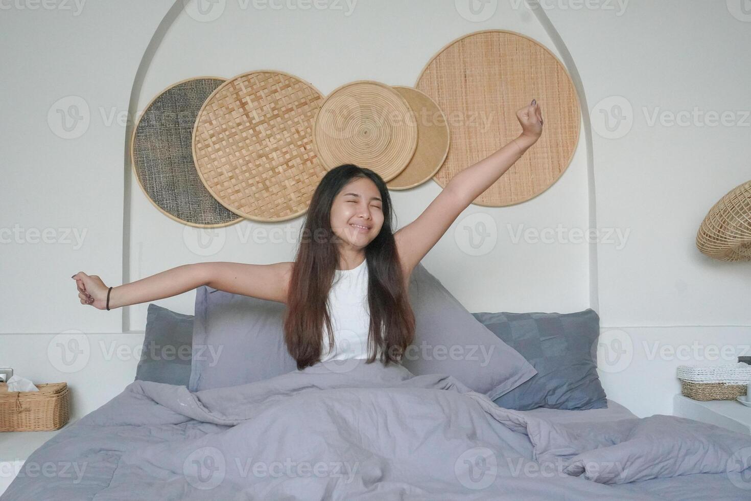 Beautiful Asian woman waking up from sleep lying on bed in bedroom, stretching hands and smiling happily photo