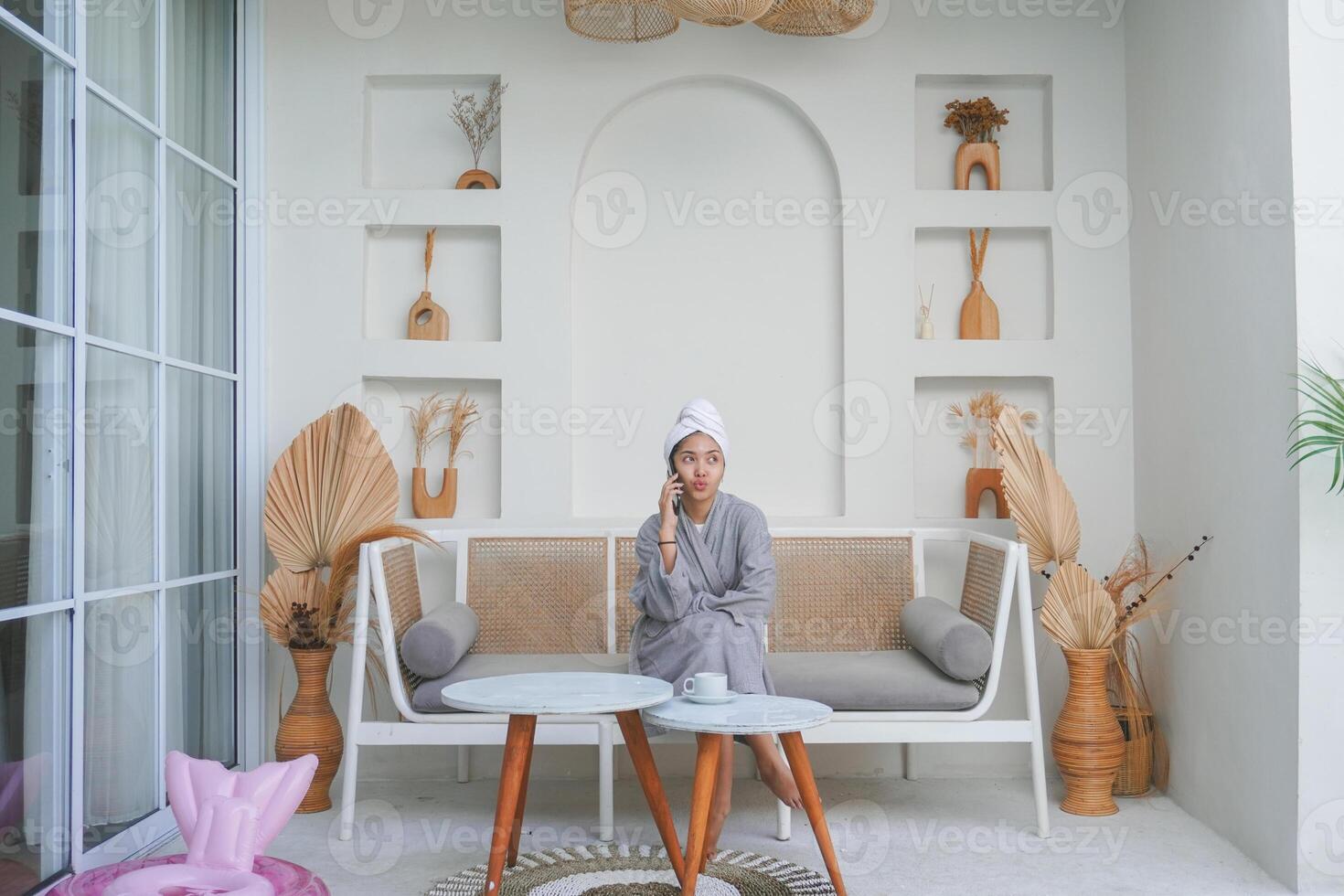 Thoughtful Asian girl in grey bathrobe sitting on a couch while thinking and having conversation on a phone call photo