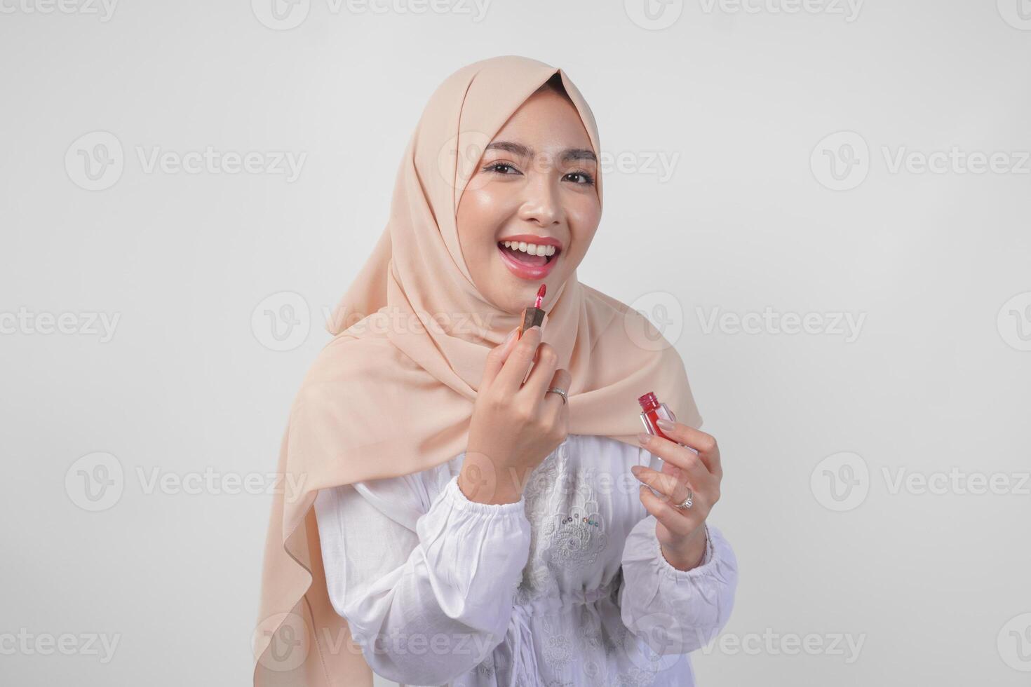 Beautiful young Asian Muslim woman wearing white dress and hijab putting on makeup applying lipstick. Fashion and cosmetics beauty concept photo