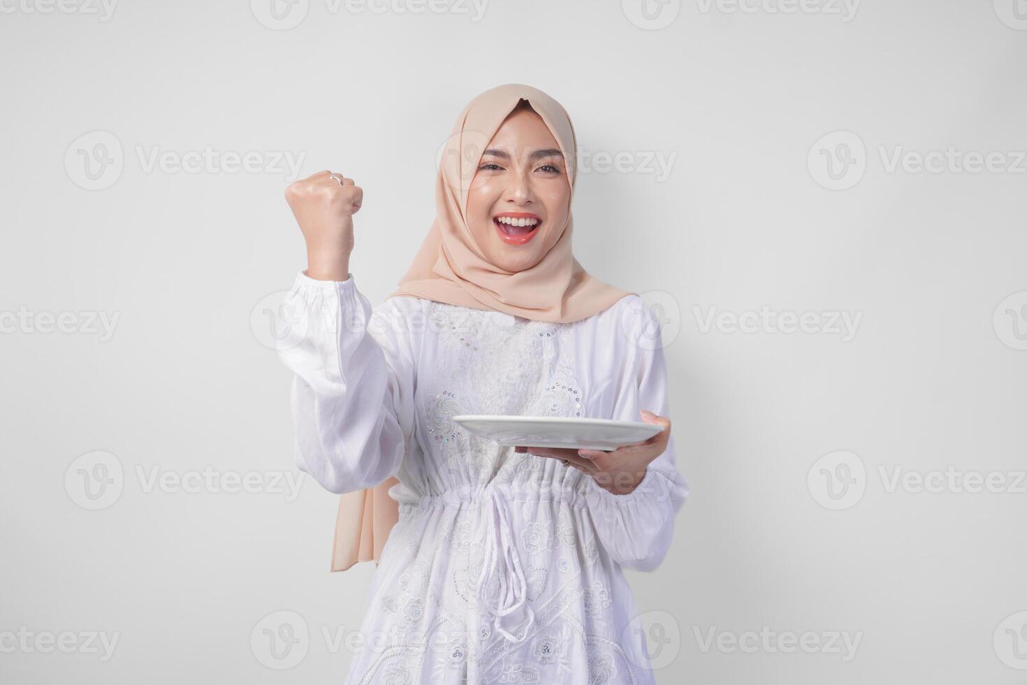 alegre joven asiático musulmán mujer en hijab haciendo un exitoso ganar gesto con apretado puño mientras presentación un vacío plato y Copiar espacio terminado eso foto