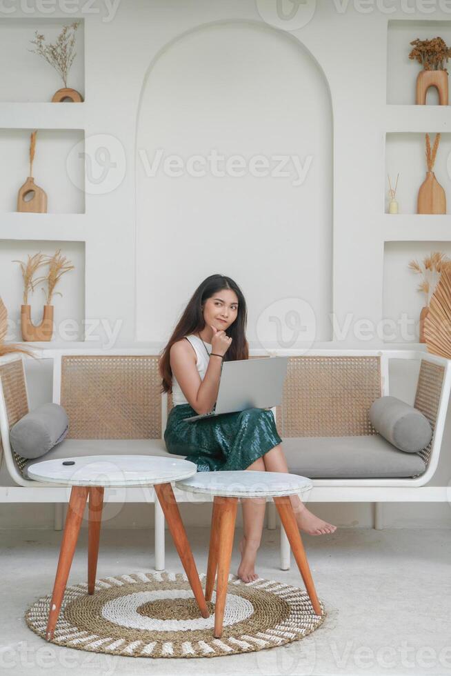 Thoughtful young woman using laptop while sitting on a sofa focused and in deep thought, working on holiday photo