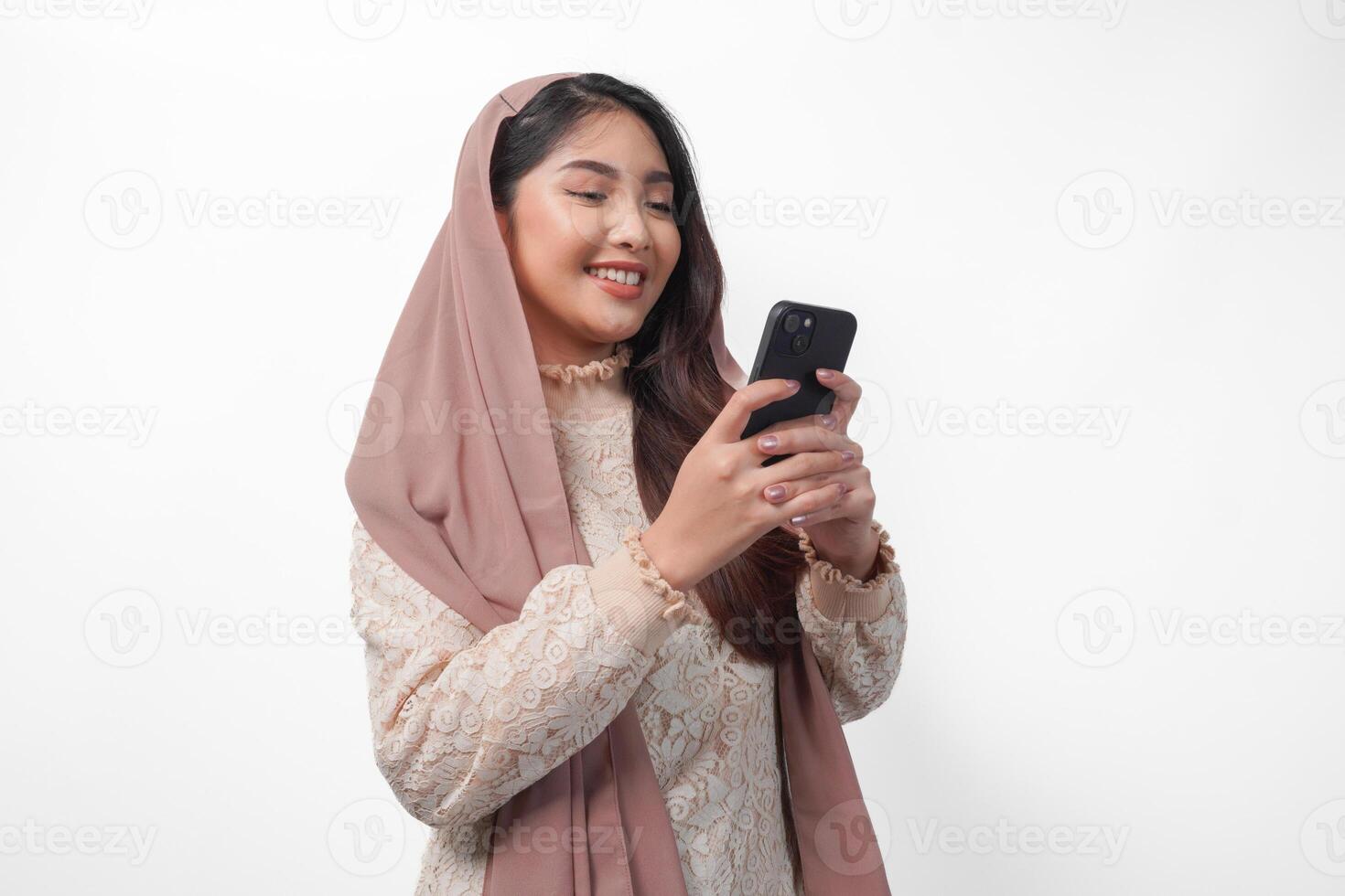 Cheerful young Asian Muslim woman in veil hijab smiling and feeling happy while holding a smartphone from side view. Ramadan and Eid Mubarak concept photo