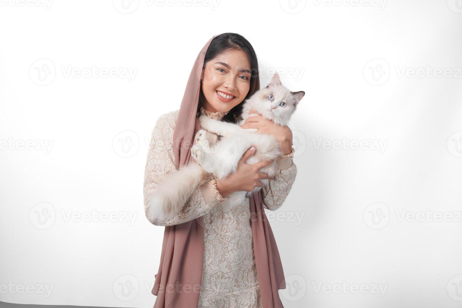 atractivo joven asiático musulmán mujer en velo hijab sonriente mientras posando y abrazando un blanco muñeca de trapo gato mascota foto