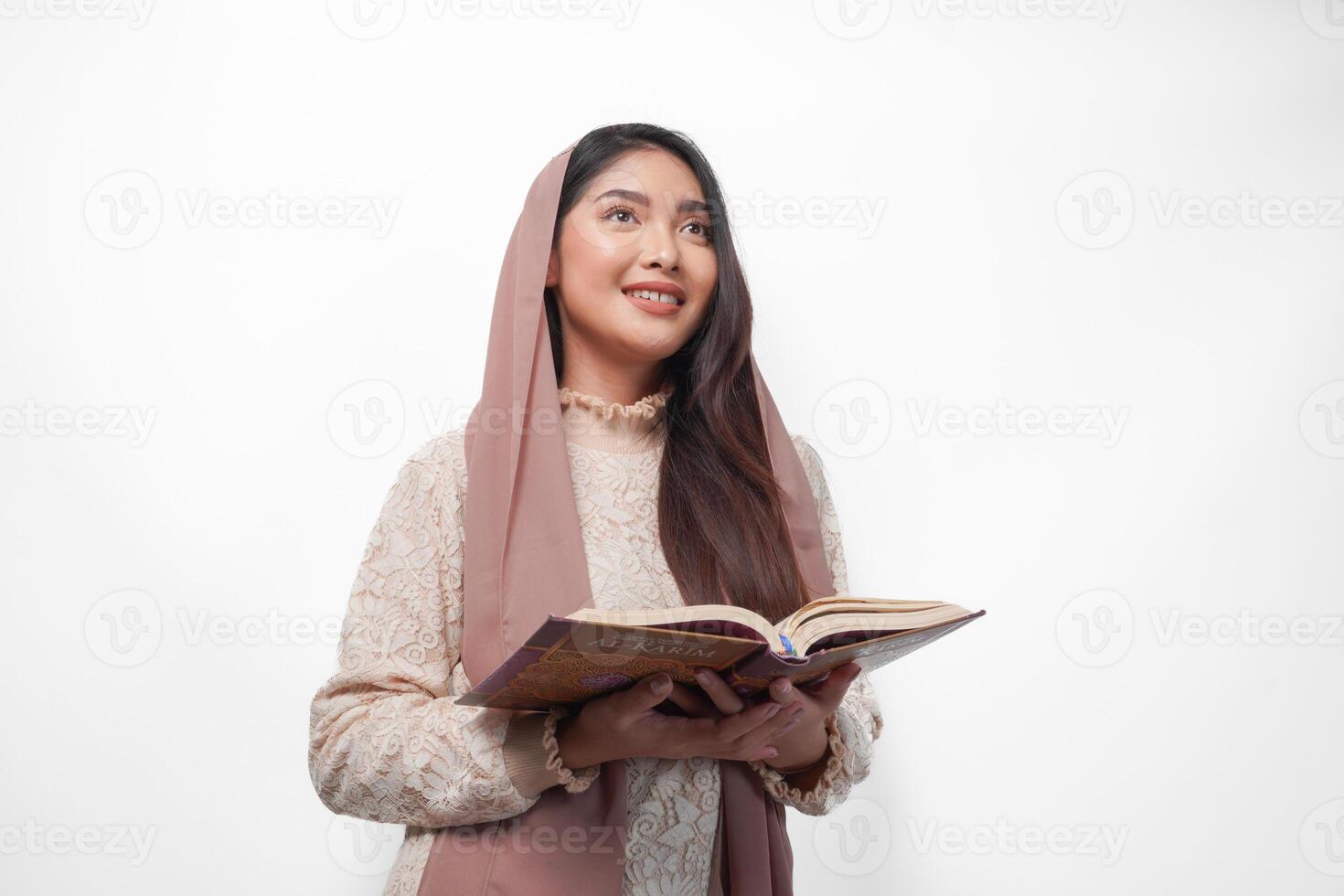 Serious Asian Muslim woman wearing veil hijab praying and reading Al Quran, standing over isolated white background. Ramadan and Eid Mubarak concept photo