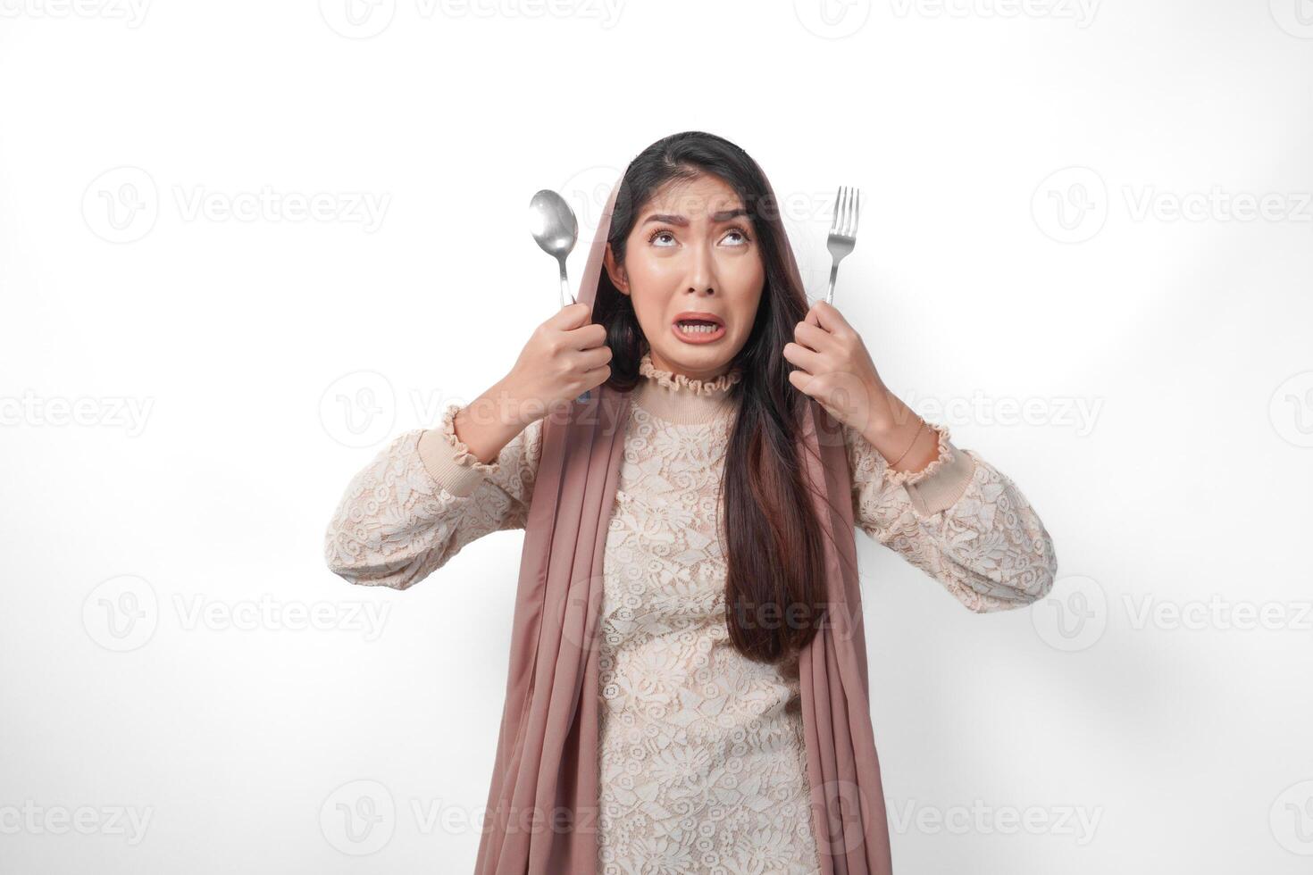 An unhappy Asian Muslim woman holding cutlery on both hands feeling stressed and frustrated, standing on isolated white background photo