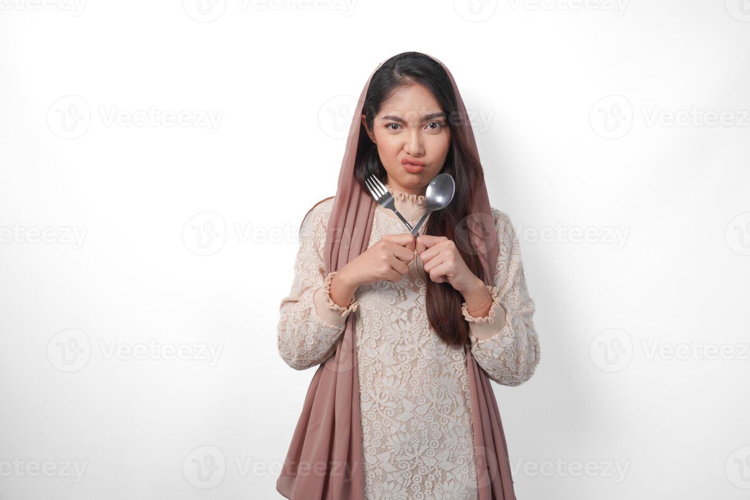 Beautiful Asian woman in veil hijab holding eating cutlery and making a refusal or rejection sign, saying no, asking to stop, standing over isolated white background photo