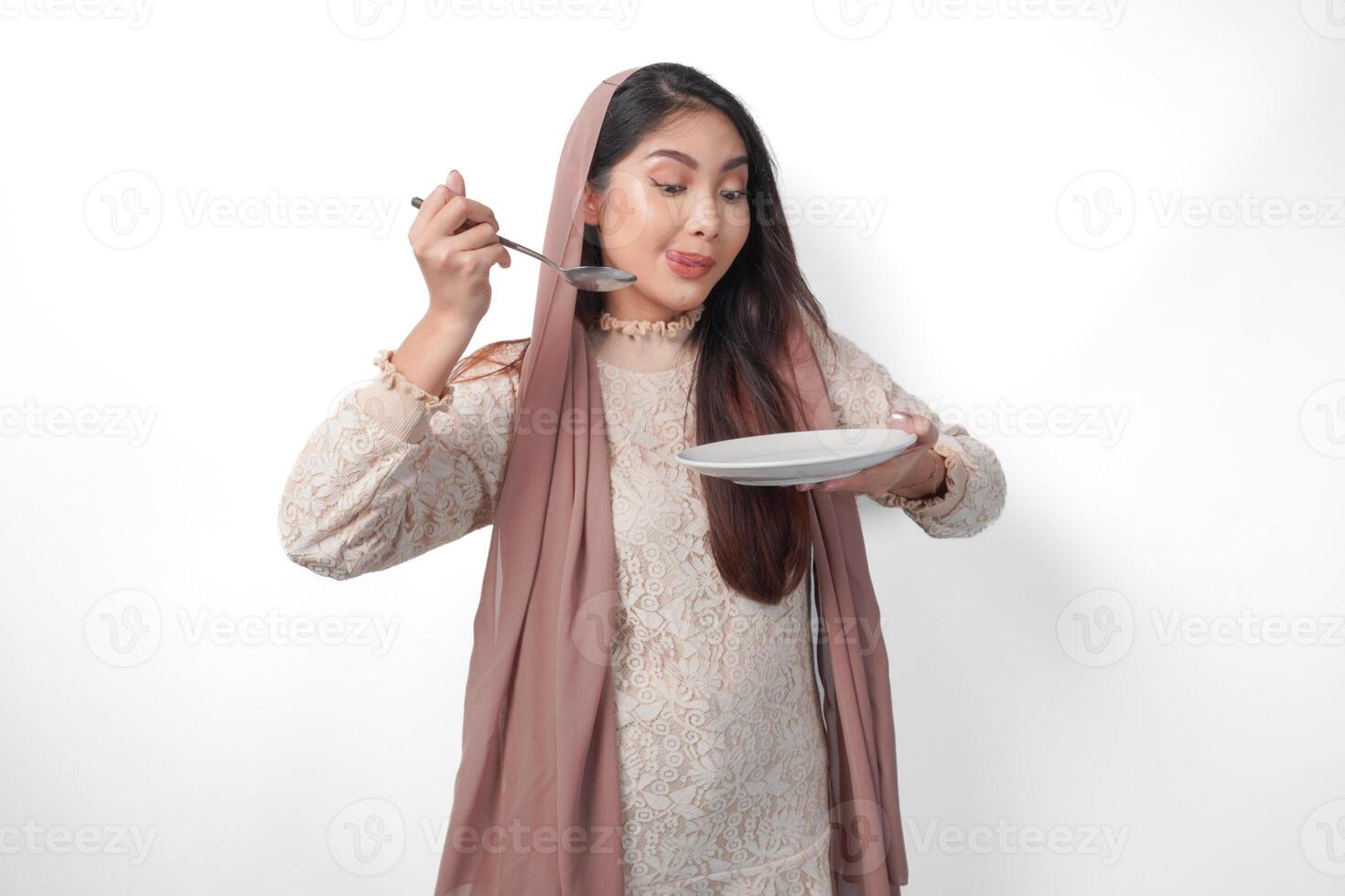 Portrait of a hungry Asian Muslim woman holding cutlery to eat or taste delicious dishes on the copy space over the plate, isolated white background photo