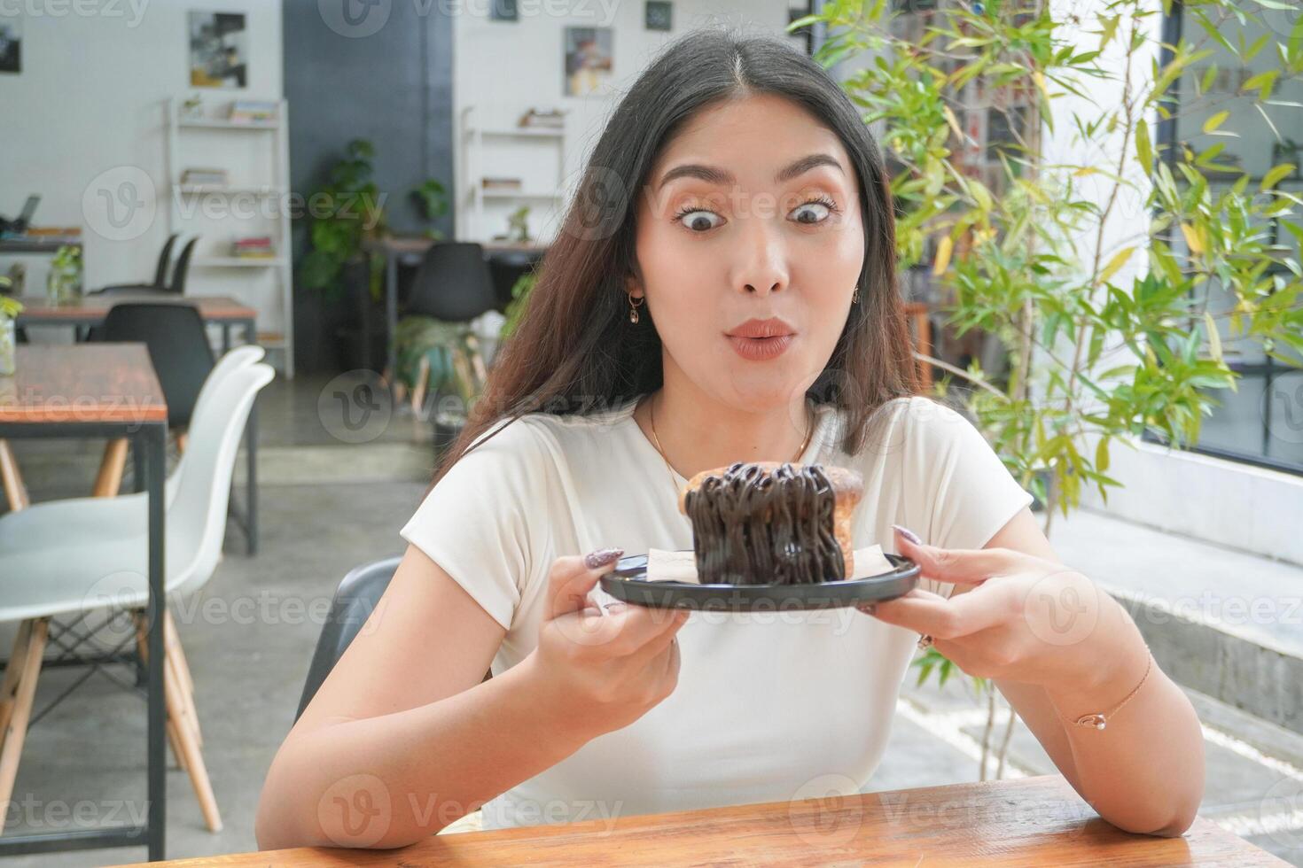 Surprised Asian young woman holding dougnut pastry named cromboloni with shocked and amazed expression, sitting in a coffeeshop with plants decoration photo