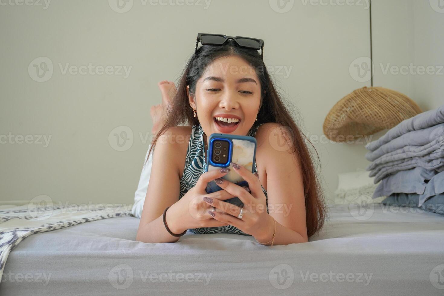 Smiling Asian woman laying down on bed with sunglasses on top while using mobile phone, exploring social media in the morning photo