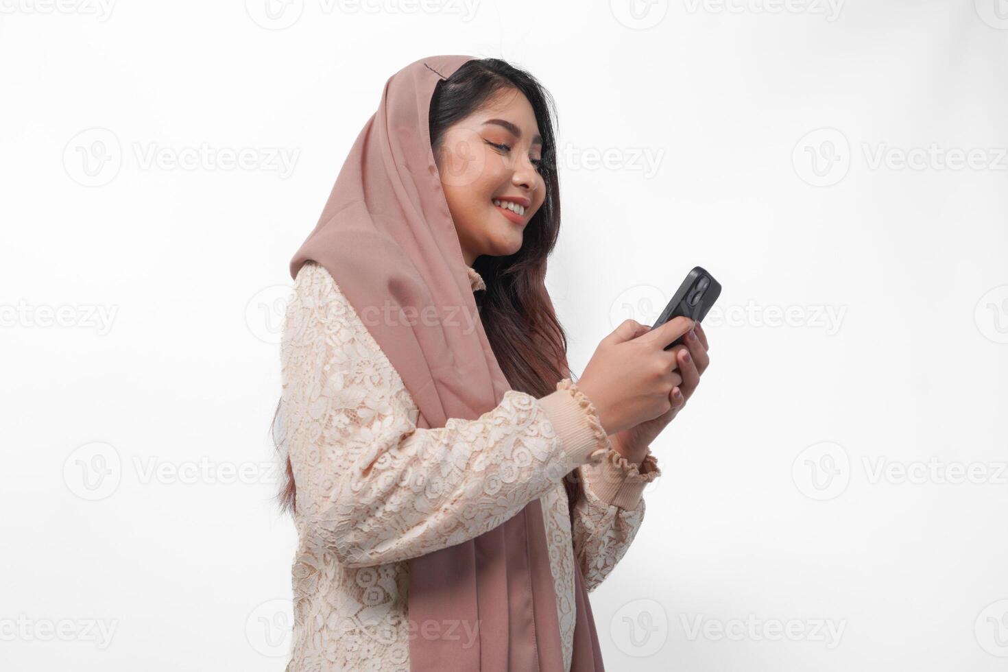 Cheerful young Asian Muslim woman in veil hijab smiling and feeling happy while holding a smartphone from side view. Ramadan and Eid Mubarak concept photo