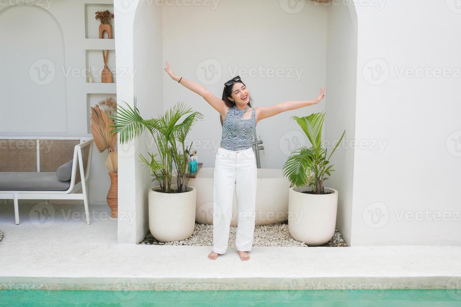 Full body of Asian young woman in pattern top and white pants spreading arms feeling excited while standing on the pool side, enjoying summer holiday photo