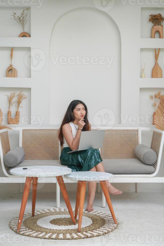 Thoughtful young woman using laptop while sitting on a sofa focused and in deep thought, working on holiday photo