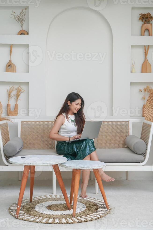 Smiling young woman in casual wear using laptop while sitting on a sofa and doing remote work on holiday as freelancer photo