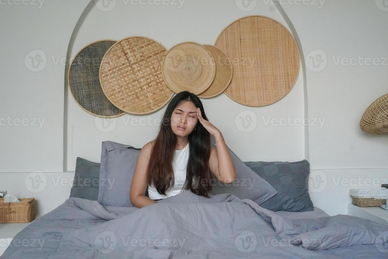 asiático mujer despertar arriba en el Mañana sensación estresado y consiguiendo dolor de cabeza después profundo dormir, todavía sensación cansado y soñoliento permanecer sentado en cama en el dormitorio foto