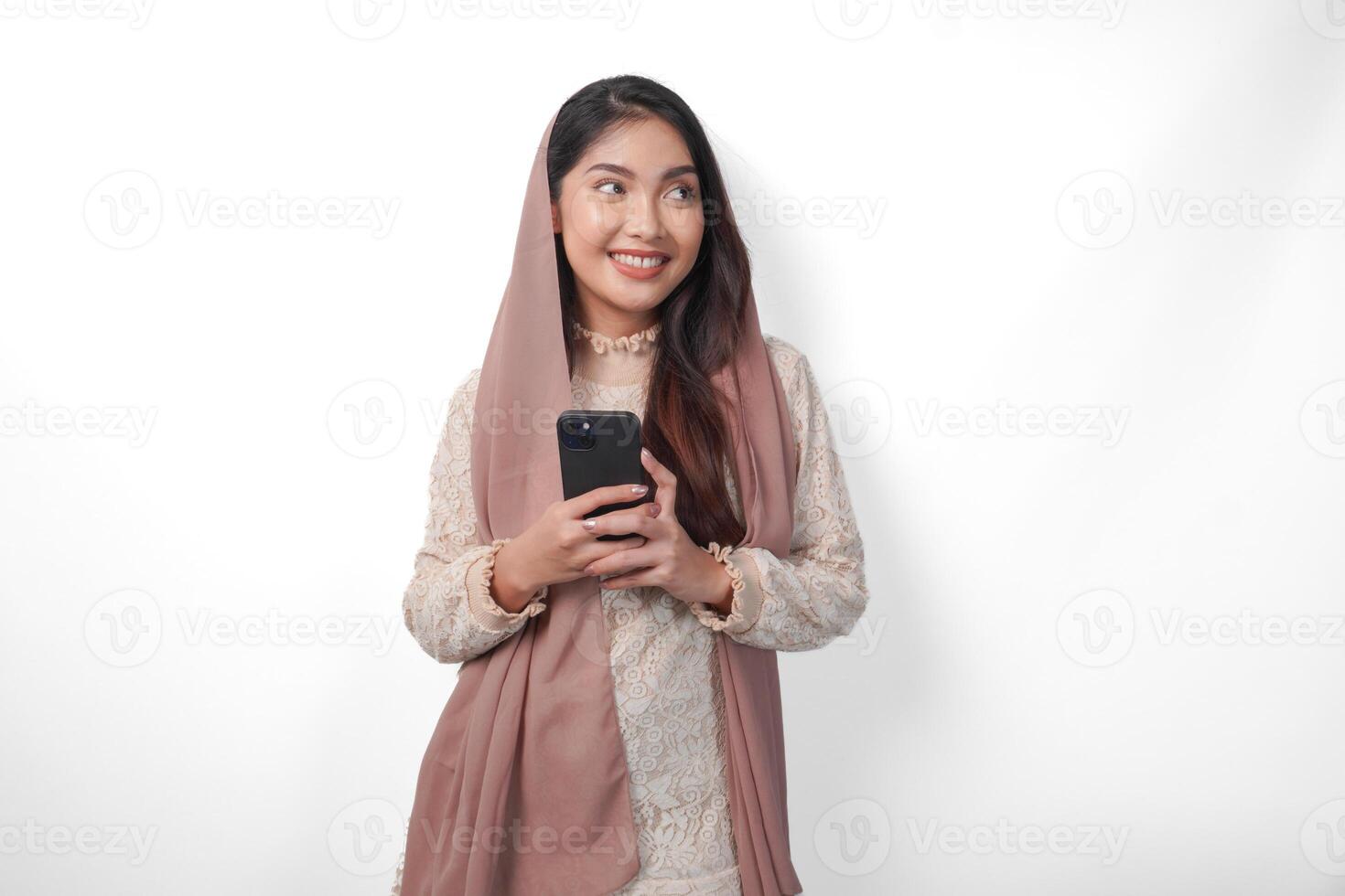 Young Asian Muslim woman in headscarf veil hijab smiling while holding a smartphone, standing over isolated white background. Ramadan concept. photo