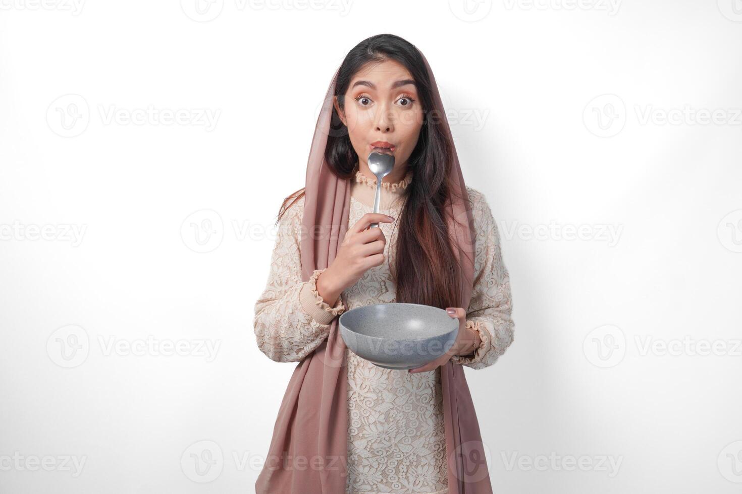 Hungry Asian Muslim woman holding bowl thinking what to eat with copy space over the bowl while biting spoon waiting for iftar, isolated white background photo