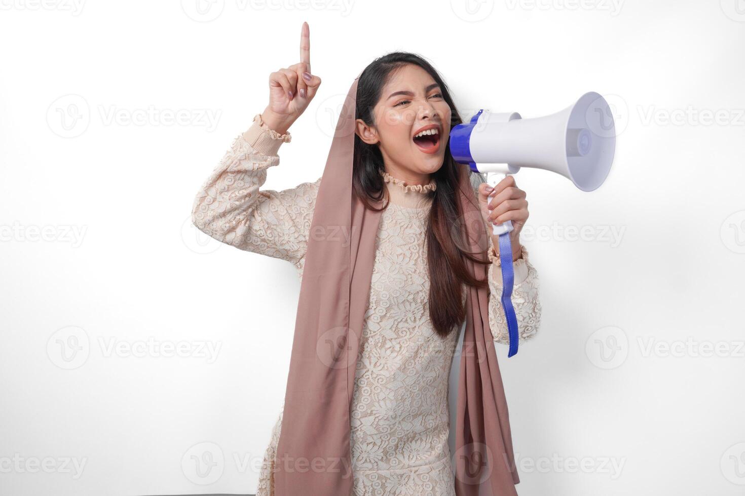 Young Asian muslim woman wearing headscarf veil hijab shouting at megaphone while pointing up to the copy space upwards, isolated on white background studio. Ramadan and Eid Mubarak concept. photo