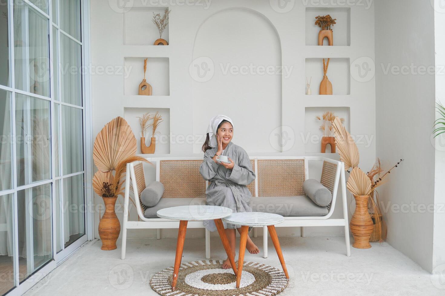 Happy attractive Asian woman in grey bathrobe posing at the terrace, smiling at camera while drinking tea. Holiday leisure concept. photo