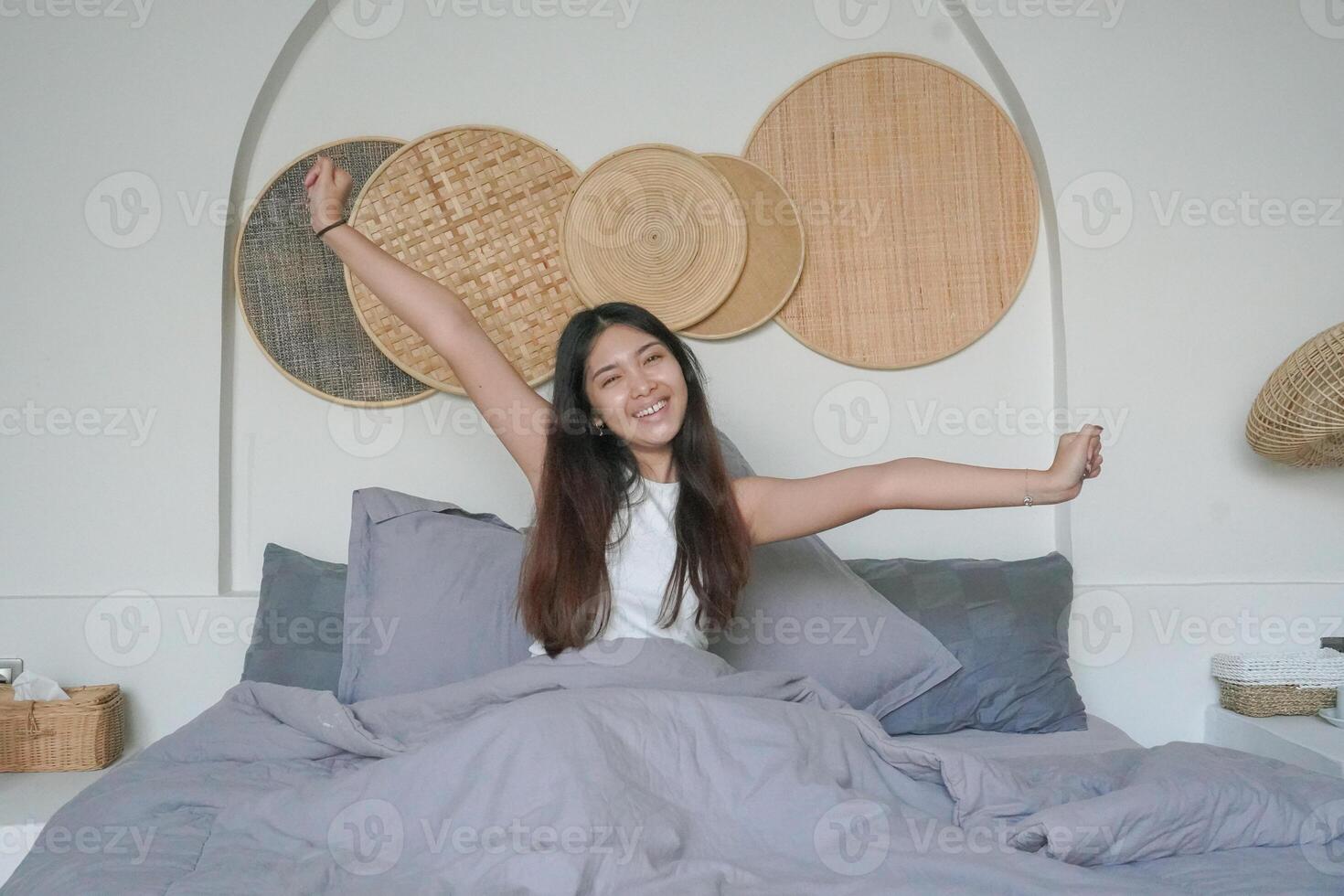 Beautiful Asian woman waking up from sleep lying on bed in bedroom, stretching hands and smiling happily photo