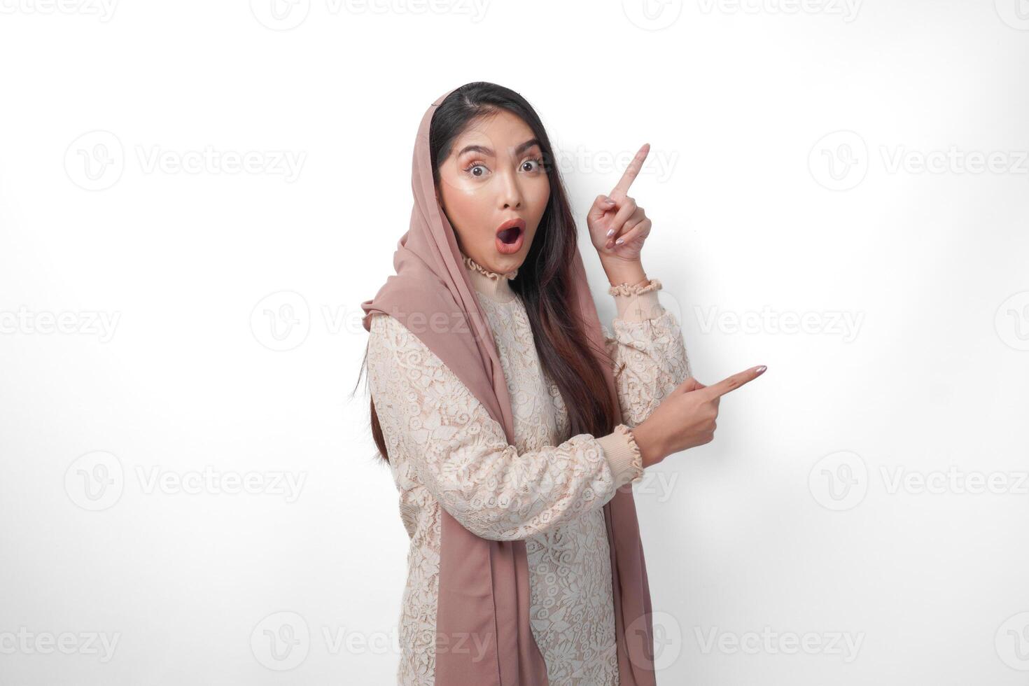 sorprendido asiático musulmán mujer vistiendo Pañuelo velo hijab mirando conmoción mientras señalando a Copiar espacio junto a su, en pie en aislado blanco antecedentes foto