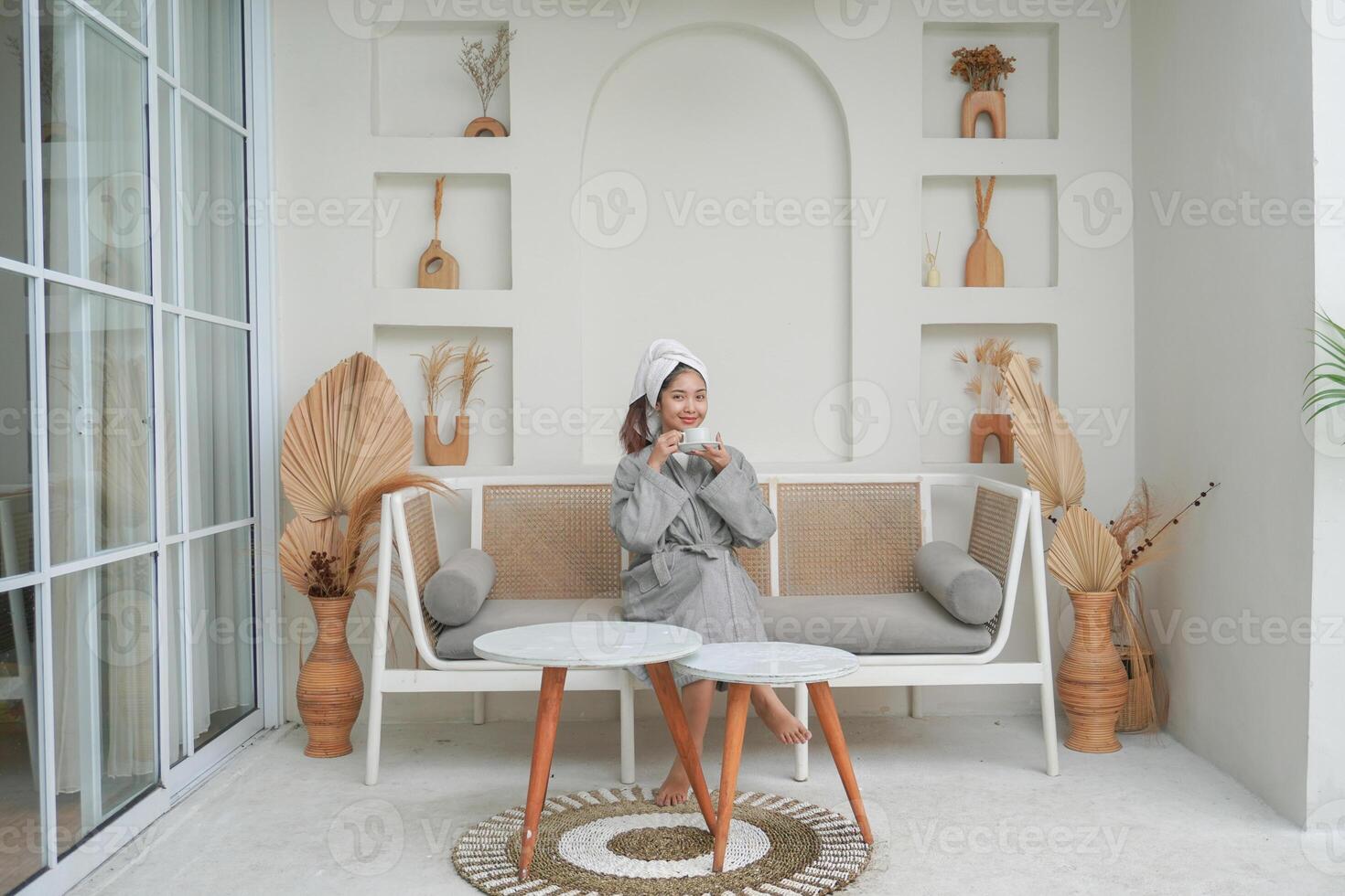 Happy attractive Asian woman in grey bathrobe posing at the terrace, smiling at camera while drinking tea. Holiday leisure concept. photo