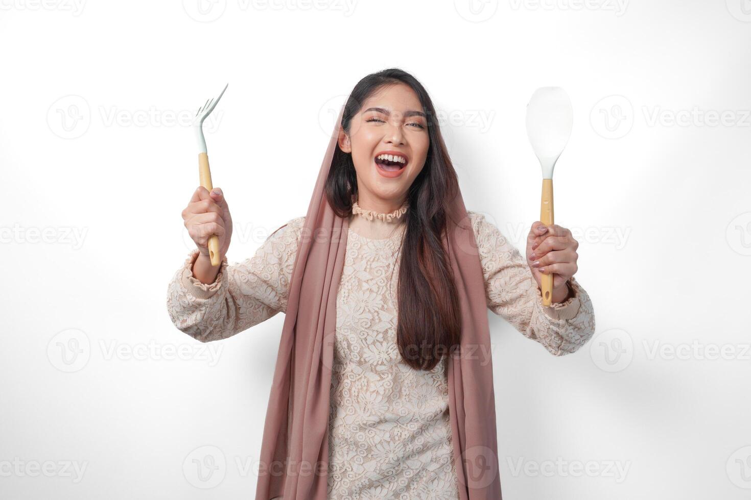 Portrait of Asian Muslim woman in veil hijab holding cooking tools or utensils while smiling cheerfully over isolated white background. Ramadan and Eid Mubarak concept photo