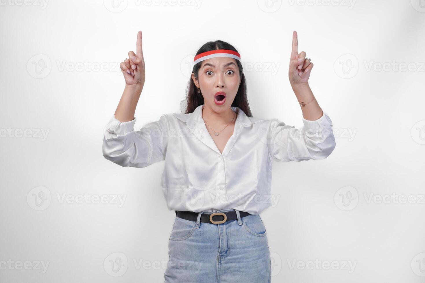 sorprendido asiático mujer vistiendo Indonesia bandera de colores venda y casual atuendo mientras señalando arriba el Copiar espacio hacia arriba. independencia día anuncio concepto foto