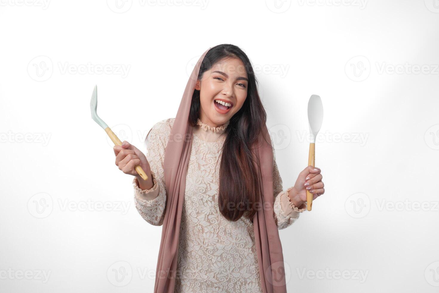 Portrait of a happy Asian Muslim woman holding kitchen cooking utensils while smiling cheerfully on isolated white background. Ramadan concept photo