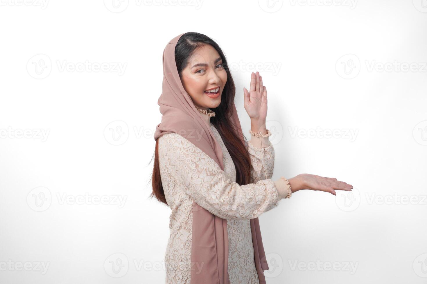 Asian Muslim woman wearing headscarf veil hijab smiling cheerfully while looking and pointing to copy space beside her, standing on isolated white background photo