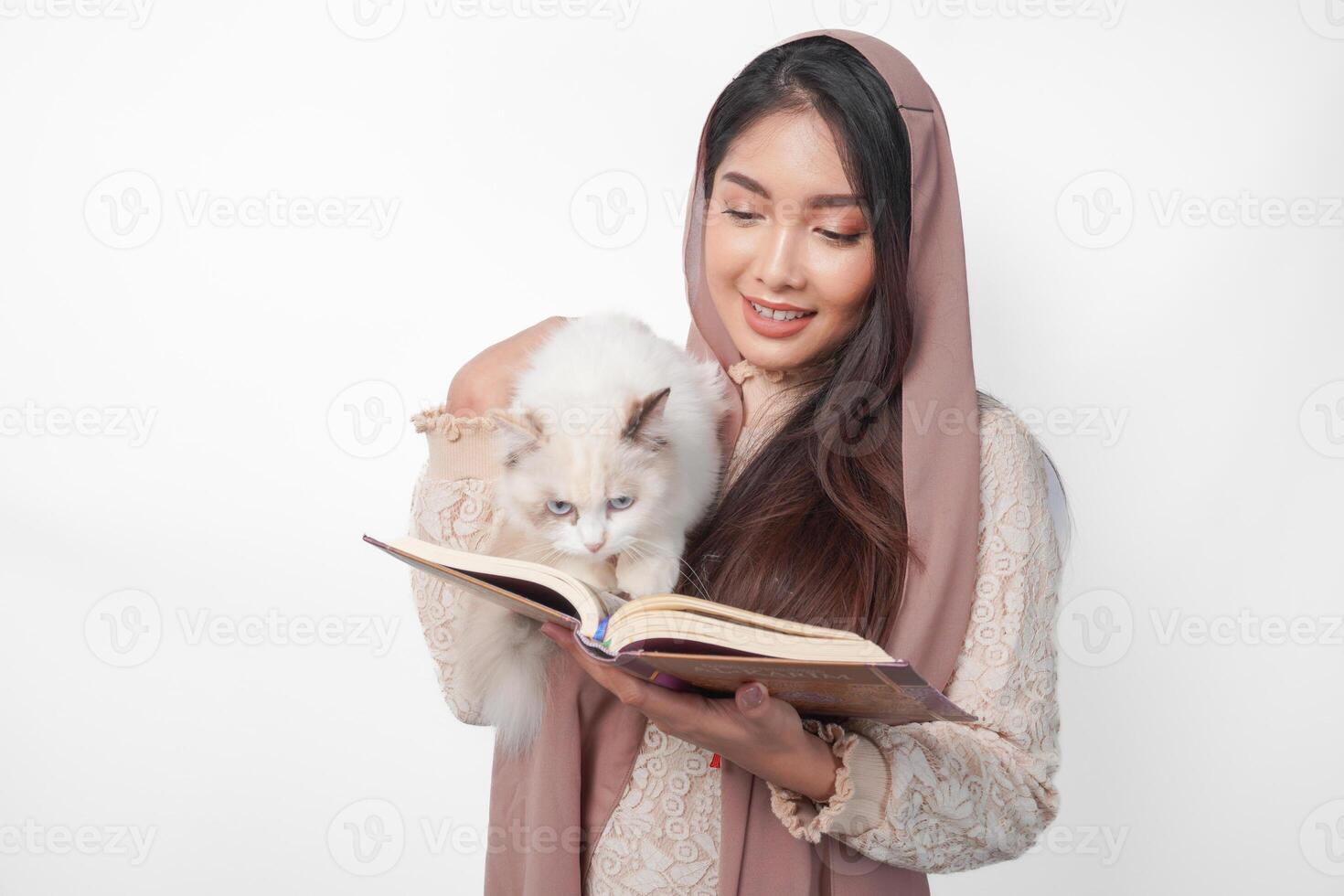 atractivo joven asiático musulmán mujer en velo hijab sonriente mientras abrazando un blanco muñeca de trapo gato mascota y participación Alabama Corán en el otro mano foto