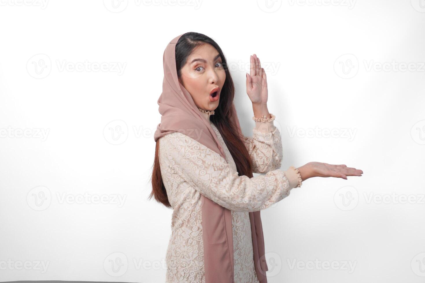 sorprendido asiático musulmán mujer vistiendo Pañuelo velo hijab mirando conmoción mientras señalando a Copiar espacio junto a su, en pie en aislado blanco antecedentes foto