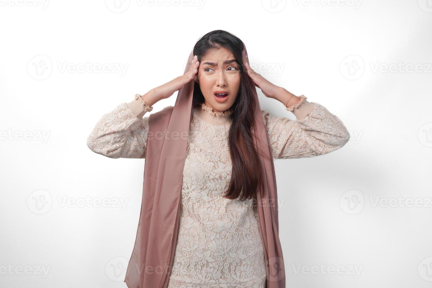 An Asian Muslim woman raising both hands to hold her head feeling stressed and overwhelmed, crying and screaming desperately, isolated white background photo